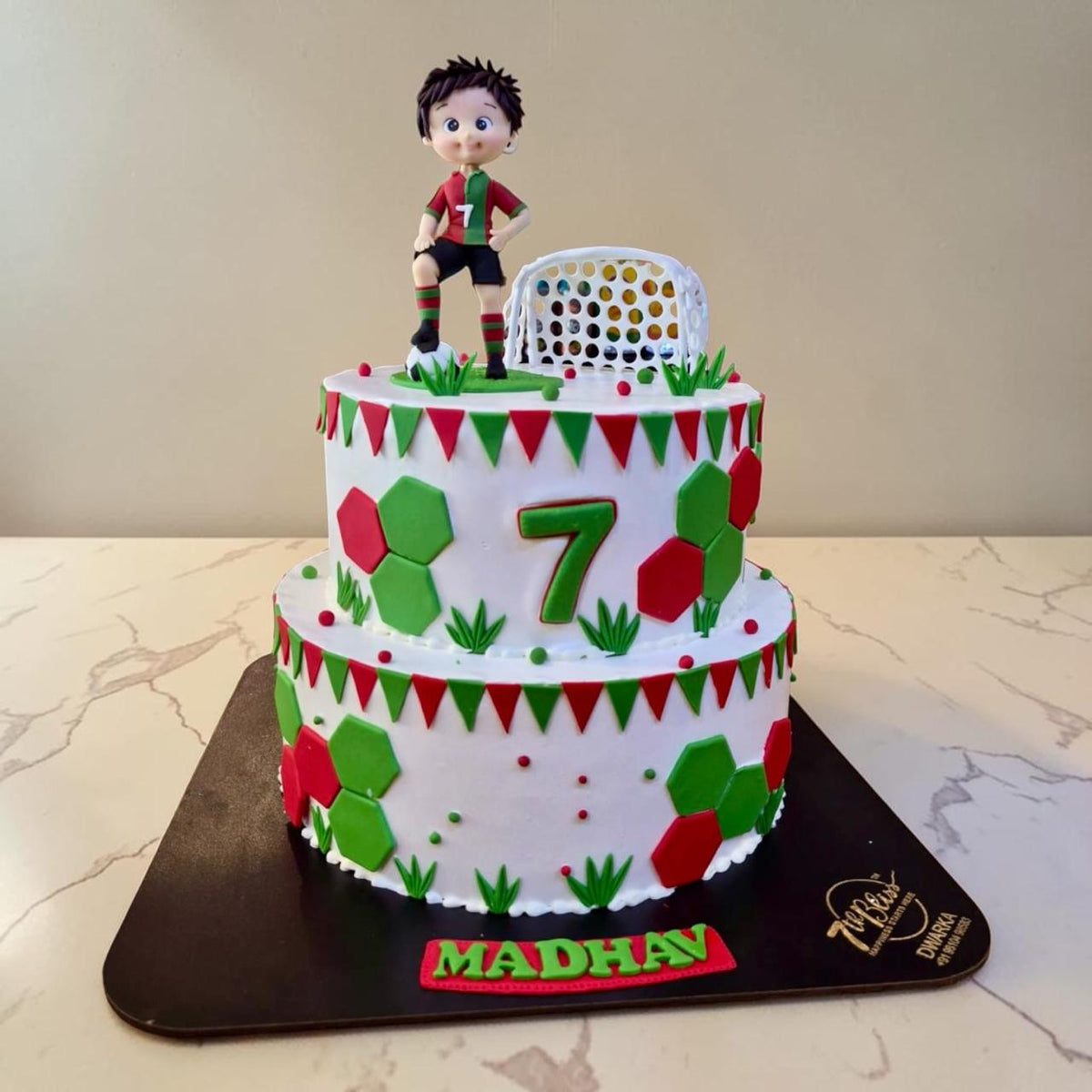 Eggless two-tier football theme birthday cake in red and green with bunting, hexagon panels and a footballer topper, 7th Bliss Dwarka Delhi